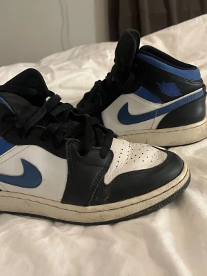 Nike Air Jordan 1 svart/blå sneakers - Säljer ett par Nike Air Jordan 1 sneakers i svart, blått och vitt. Klassisk high-top modell med snörning och rund tå. Skorna har blå swoosh och Air Jordan-logga på sidan, tillverkade i skinn med perforerad tåbox för extra ventilation.
