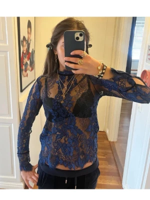 Blå spetsblus med lång ärm - Säljer en transparent blå blus i spets med blommigt mönster och lång ärm. Blusen har hög hals och är figurnära, perfekt för att styla med smycken och lager på lager. Supersnygg att bära över ett linne eller bh.