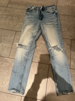 Blå ripped jeans från Zara, strl 34 - Säljer ett par blå jeans från Zara i storlek 34 med snygga slitningar och hål vid knäna. Jeansen har klassisk femficksdesign, dragkedja och knapp framtill. Materialet är jeans och passformen är rak med lite loose vibe.
