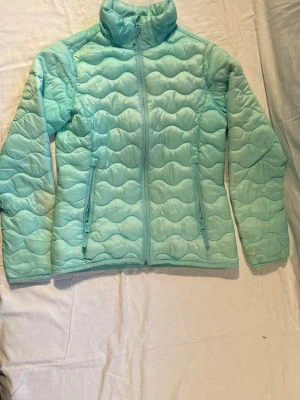 Ljusblå McKinley pufferjacka 158 - Snygg ljusblå pufferjacka från McKinley i storlek 158. Jackan har vågquiltat mönster, hög krage och dragkedja framtill samt två sidofickor med dragkedja. Perfekt för kyliga dagar och enkel att matcha med din vardagsstil.