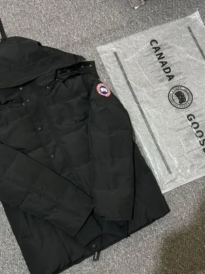 Svart macmillan jacka från Canada Goose XL - Säljer en svart dunjacka från Canada Goose i storlek XL. Jackan har huva, klassisk patch på ärmen och stängs med både dragkedja och knappar. Perfekt för kalla dagar med sin rejäla vaddering och stilrena look.
