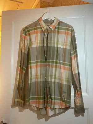Rutig skjorta från Acne i storlek 50 - Säljer en snygg rutig skjorta från Acne i storlek 50. Skjortan har ett färgglatt rutmönster i grönt, orange, beige och vitt, klassisk krage och knappar framtill. Tillverkad i mjuk bomull och har långa ärmar. Perfekt för dig som gillar stilrena och coola plagg.