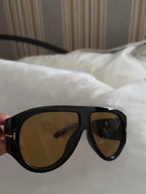 Svarta Tom Ford modell pilotbågar solglasögon - Snygga solglasögon med svart, bred båge och bruna tonade glas. Klassisk pilotform med guldfärgade detaljer vid gångjärnen. Perfekta för dig som gillar en statement-look och vill ha något trendigt och exklusivt.