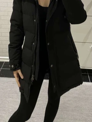 Svart lång dunjacka med dragkedja - Snygg svart dunjacka med lång passform och dragkedja framtill. Jackan har quiltad design och långa ärmar, perfekt för kalla dagar. Enkel och stilren look som passar till det mesta.