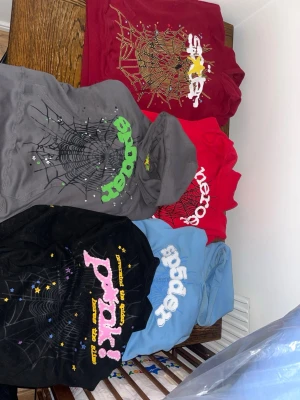 Färgglada hoodies från Sp5der - Säljer flera hoodies från Sp5der i olika färger: svart, grå, ljusblå, röd och vinröd. Alla har coolt spindelnätsmönster och färgglada tryck på bröstet. Hoodiesarna har huva och en avslappnad passform, perfekta för en streetwear-look.