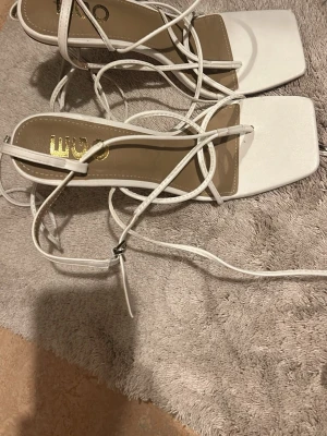 Vita sandaler med snörning och klack - Säljer ett par vita klackskor med smala remmar och snörning runt vristen. Skorna har fyrkantig tå och genomskinlig hög klack som ger en modern vibe. Tillverkade i syntetmaterial med stilren look, perfekta för att lyfta din outfit. Storlek 39. Använt 1 gång.