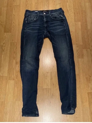 Mörkblå Replay Anbass jeans - Säljer ett par mörkblå Replay Anbass jeans med snygga slitningar och klassisk femficksdesign. Jeansen har smal passform och är tillverkade i stretchig bomullsdenim för extra komfort. Perfekta för dig som gillar en modern och stilren look. Storlek 30/32