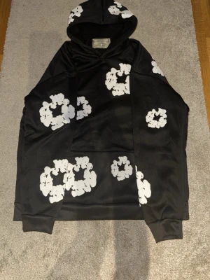 Svart hoodie från Denim Tears med blommor - Svart hoodie från Denim Tears med vita blommönster över hela tröjan och på huvan. Hoodien har en klassisk passform med ribbade muddar och stor känguruficka framtill. Perfekt för dig som gillar streetwear och vill sticka ut med en unik design.