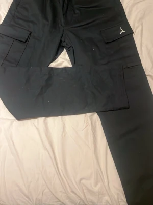 Svarta byxor från maniére de voir helt nya - Säljer ett par svarta cargopants med stora sidofickor och en snygg detalj i form av en vit logga på ena ficklocket. Byxorna har rak passform och är perfekta för en avslappnad streetwear-look. Materialet känns slitstarkt och bekvämt.
