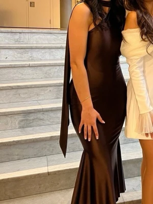 Oh Polly Brun asymmetrisk satinklänning  - Snygg brun satinklänning med asymmetrisk axel och lång scarfdetalj. Klänningen har figurnära passform och lätt utsvängd nederdel. Perfekt för dig som vill sticka ut med en elegant och modern look. Använd endast en gång, tvättad och ren. 