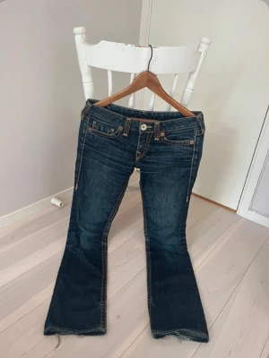 Mörkblå bootcut jeans med broderade fickor - Snygga true religion jeans med bootcut-modell och coola broderade bakfickor. Klassisk femficksdesign, orangea sömmar och låg midja. Perfekta för dig som gillar en avslappnad men trendig stil. Små hål ena på insidan av benet och den andra på sidan av knäet (se bild 3)
