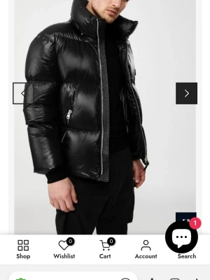 Svart dunjacka från Mackage - Stilren svart dunjacka från Mackage med glansig finish, hög krage och dragkedja framtill. Jackan har två fickor med dragkedja och tydlig logotyp på ärmen. Perfekt för kalla dagar när du vill hålla värmen och se cool ut.