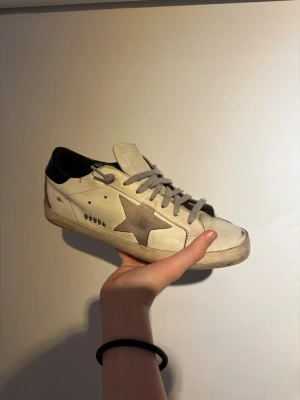 Golden goose - Golden Goose sneakers i vitt skinn med grå snörning och en grå stjärna på sidan. Skon har svart hälparti och klassisk låg siluett. Snygg vintage-look och platt sula. Perfekt för dig som gillar streetstyle och unika detaljer.