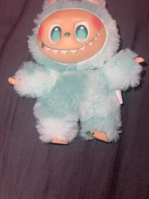 Labubu - Unik och gullig monsterdocka med stort ansikte, glittriga gröna ögon och ett brett leende med vita tänder. Dockan har en mjuk, ljusblå fluffig kropp och små öron. Perfekt som prydnad eller samlarobjekt för dig som gillar söta och annorlunda figurer.