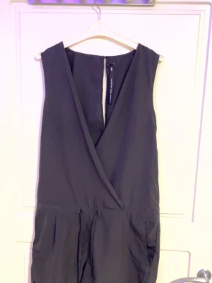 Svart omlott jumpsuit från Monki - Snygg svart ärmlös jumpsuit från Monki med omlott framtill och djup v-ringning. Modellen har fickor i sidorna och en liten knapp i nacken för stängning. Perfekt för dig som gillar en stilren och modern look.