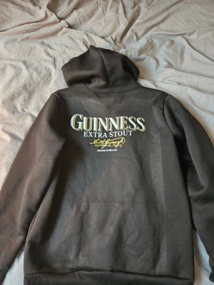Svart Guinness hoodie - rygg tryck - Svart hoodie med huva och magficka från Guinness. Riktigt fett backprint med den klassiska "Extra stout" på. Perfekt för dig som gillar guiness