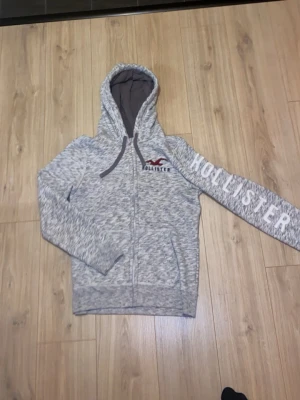 Grå zip hoodie från Hollister - Snygg gråmelerad hoodie från Hollister med dragkedja framtill och stor huva. Logga på bröstet och stort HOLLISTER-tryck längs ena ärmen. Mjukt material och fickor på sidorna. Perfekt för en avslappnad och trendig look.