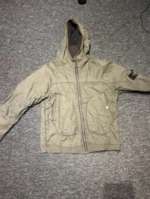 Beige vindjacka från Stone Island - Snygg beige vindjacka från Stone Island med huva och ikonisk patch på ärmen. Jackan har två stora fickor framtill och dragkedja. Perfekt för dig som vill ha en stilren och funktionell jacka med streetwear-vibe.