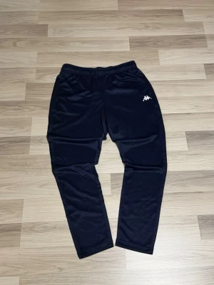 Mörkblåa mjukisbyxor från Kappa - Säljer ett par mörkblåa mjukisbyxor från Kappa med klassisk logga på vänster lår. Byxorna har elastisk midja och smal passform, perfekta för chill eller träning. Tillverkade i mjukt och lätt polyester-material.