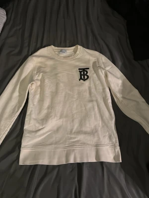 Vit sweatshirt från Burberry - Säljer en stilren vit sweatshirt från Burberry med det klassiska TB-logotypen tryckt i svart på bröstet. Tröjan har rund halsringning och långa ärmar. Fick den i present för några år sedan och har sedan desss blivit för liten. Använt den 3-4 gånger totalt 