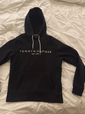Svart hoodie från Tommy Hilfiger - Snygg svart hoodie från Tommy Hilfiger med vit snörning i huvan och klassisk logga framtill. Hoodien har känguruficka och mjukt material som passar perfekt till en avslappnad stil.