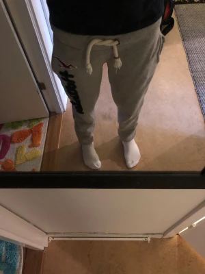 Gråa mjukisbyxor från Hollister - Gråa mjukisbyxor från Hollister med svart broderad logga längs benet och vita snören längst upp. Byxorna har sidofickor. Perfekta för chill dagar och riktigt sköna tack vare mjukt bomullsmaterial. En måste i garderoben! 