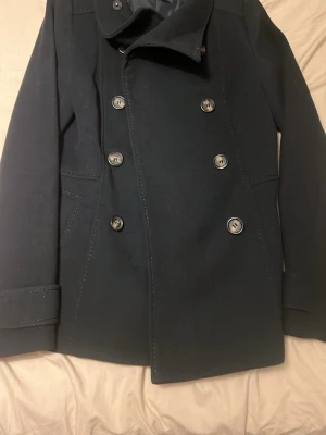 Mörkblå skepparkavaj med knappar storlek:34 - Stilren mörkblå skepparkavaj med dubbelknäppning och stora silvriga knappar. Klassisk krage och två fickor framtill. Perfekt för dig som vill ha en tidlös och snygg jacka till höst och vinter.