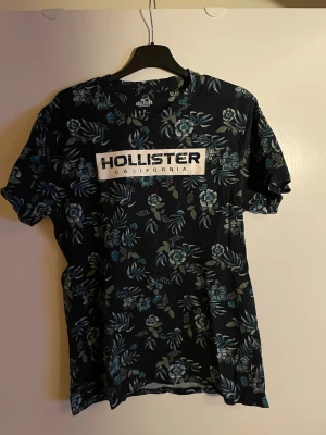 Blommig Hollister t-shirt marinblå - Snygg marinblå t-shirt från Hollister med stort tryck fram och bak. T-shirten har blommigt mönster i blå och gröna toner samt klassisk rund hals och korta ärmar. Tillverkad i mjuk bomull som känns skön mot huden. Perfekt för dig som gillar streetwear och tydliga loggor.