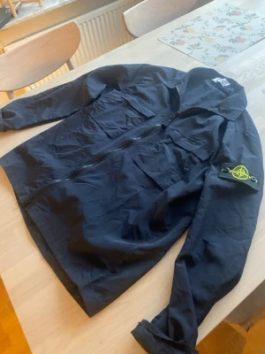 Marinblå overshirt Stone Island - Säljer en svart overshirt från Stone Island med klassisk patch på ärmen. Jackan har två bröstfickor, dragkedja framtill. Perfekt för lager-på-lager och streetwear-stil. Hör av dig vid frågor elr liknande!