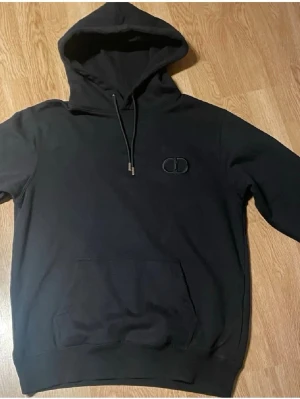 Svart hoodie f - Snygg svart hoodie från med broderad CD-logga på bröstet. Klassisk modell med huva, dragsnören och stor magficka framtill. Perfekt för en avslappnad och trendig look.