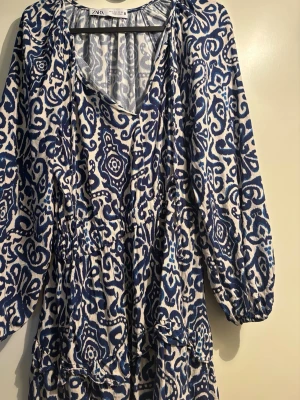 Blåvit mönstrad klänning från Zara - Säljer en blåvit klänning från Zara med v-ringning och lång ärm. Klänningen har ett unikt abstrakt mönster, resår i midjan och volangdetaljer. Tillverkad i ett lätt och luftigt material, perfekt för varmare dagar.