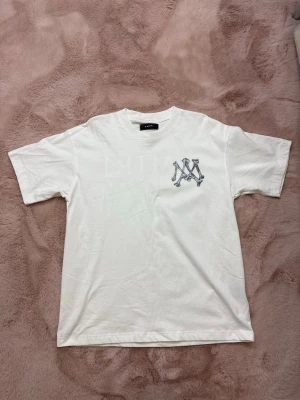 Amiri T-Shirt - Vit t-shirt från Amiri med ett coolt beninspirerat logotryck på bröstet. Klassisk rund halsringning och korta ärmar. Perfekt för dig som gillar streetwear och vill ha något stilrent men ändå unikt.