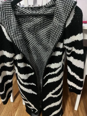 Svartvit stickad kofta med zebramönster - Cool svartvit stickad kofta med zebramönster och kontrasterande mönster vid axlarna. Koftan är lång och har öppen front, perfekt för lager-på-lager-stil. Materialet är mjukt och mysigt, passar dig som vill sticka ut.
