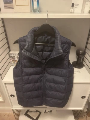 Mörkblå  dunväst från uniqlo - Säljer en mörkblå quiltad dunväst från uniqlo med hög krage och dragkedja framtill. Västen har en stilren look och är perfekt att slänga över en hoodie eller tröja för extra värme. Passar dig som gillar en enkel men snygg stil. Strl XS men kan passa S i vissa fall