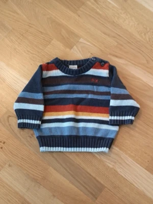 Randig stickad tröja - Mysig stickad tröja från FIX i storlek 56. Tröjan har breda ränder i blått, brunt, orange, vitt och gult samt två knappar på axeln för enkel påklädning.