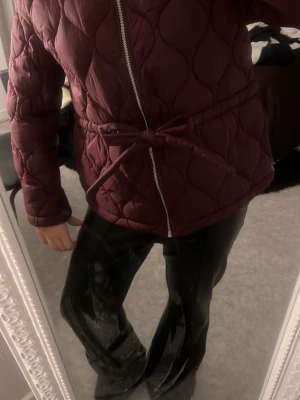 Vinröd quiltad pufferjacka med knyt - Säljer en vinröd quiltad pufferjacka med dragkedja och knyt i midjan. Jackan har lång ärm och en snygg, figurnära passform. Perfekt för kyligare dagar och ger en trendig look med sitt vågiga quiltade mönster.