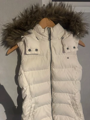 Vit dunväst från Tommy Hilfiger - Säljer min älskade vita dunväst från Tommy Hilfiger med avtagbar huva och fejkpäls. Liten logga på bröstet. Perfekt under vår, höst och vinter.