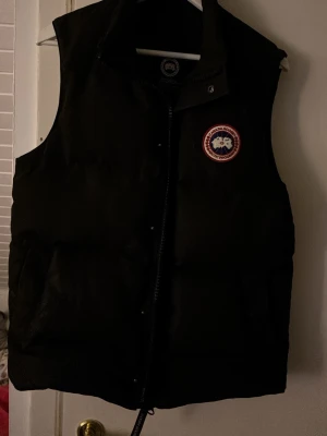 Svart dunväst från Canada Goose - Snygg svart dunväst från Canada Goose med klassisk logga på bröstet. Västen har hög krage, dragkedja och tryckknappar framtill samt två stora fickor. Perfekt för lager-på-lager och håller dig varm under kyliga dagar.