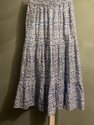 Blåvit mönstrad långkjol - Snygg långkjol med blått och vitt mönster. Kjolen har volangdetaljer och resår i midjan för en bekväm passform. Perfekt för dig som gillar en avslappnad och trendig stil. Har använt en eller två gånger.
