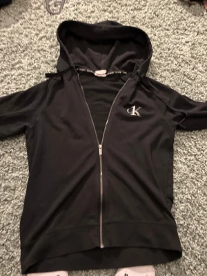 Svart hoodie från Calvin Klein - Svart zip-hoodie från Calvin Klein med dragkedja framtill och huva. Diskret CK One-logga på bröstet och Calvin Klein-tryck på insidan av huvan. Tillverkad i mjuk bomull, perfekt för en avslappnad stil.