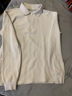 Vit långärmad pikétröja med dragkedja - Vit långärmad pikétröja med klassisk krage och dragkedja framtill. Tröjan har ribbade muddar vid ärmslut och är tillverkad i ett strukturerat tyg som ger en sportig känsla. Perfekt för en clean och stilren look.