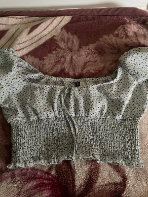 Blommig croppad blus med puffärm - Söt blommig croppad blus från H&M med puffärmar och smockad midja. Toppen har små blå och gröna blommor på vit botten, knytband framtill och volangkant nedtill. Perfekt för en trendig och somrig look.