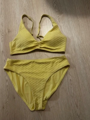 Gul bikini med struktur - Säljer en gul bikini med snygg struktur i tyget. Överdelen har breda axelband och lätt rynkning framtill, medan underdelen är högmidjad för en trendig look. Perfekt för soliga dagar på stranden eller vid poolen. Storlek S/M.