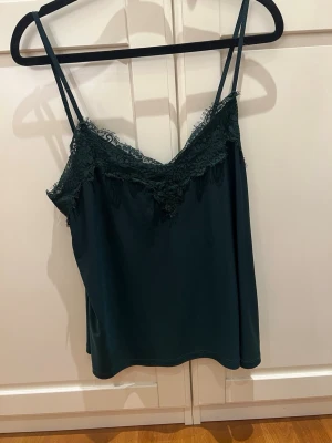 Mörkgrön linnetopp med spets - Snygg mörkgrön linnetopp med tunna axelband och dekorativ spets längs urringningen. Toppen har en lätt glansig finish och ett avslappnat fall. Perfekt att styla med jeans eller kjol för en trendig look.