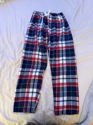 Rutig pyjamasbyxa med rosett - Mysiga pyjamasbyxor i rött, blått, vitt och grått med klassiskt rutigt mönster. Byxorna har en bred resår i midjan och en vit rosett framtill. Perfekta för chillkvällar eller lata morgnar.