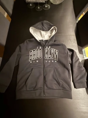 Svart zip hoodie Brooklyn från SHEIN - Snygg svart zip hoodie från SHEIN med vit text 'BROOKLYN 1998 NEW YORK' på bröstet. Hoodien har vit insida i huvan, två fickor fram och dragkedja. Perfekt för en avslappnad streetwear-look.
