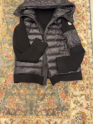 Moncler cardigan med huva - Snygg svart dunväst från Moncler med quiltad framsida, stickade svarta ärmar och breda muddar. Västen har en praktisk huva och dragkedja framtill samt en ikonisk Moncler-logga på bröstet. Perfekt för lager-på-lager och streetstyle.