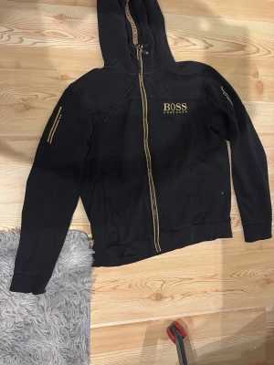 Svart hoodie från Hugo Boss - Snygg svart hoodie från Hugo Boss med guldfärgade detaljer och dragkedja. Märkets logga är broderad på bröstet i guld. Hoodien har huva med snörning och ribbade muddar. Materialet är mjukt och bekvämt, perfekt för en avslappnad stil.