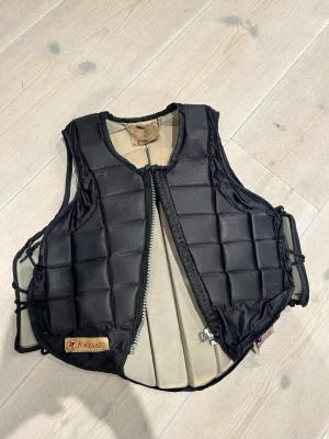 Racesafe ridväst  - Säljer en svart ridväst från Racesafe med vadderad design och dragkedja framtill. Västen har justerbara snören i sidorna och är tillverkad i slitstarkt material. Barnstorlek i M. Orginalpris: 2 949kr 