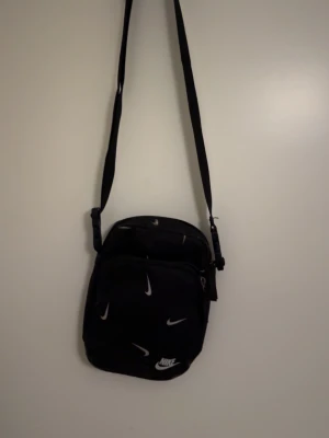 Svart axelväska från Nike - Den har använts och man ser därför priset Snygg svart axelväska från Nike med justerbar rem och flera vita swoosh-loggor över hela väskan. Väskan har dragkedja och är perfekt för att bära det viktigaste. Tillverkad i slitstarkt syntetmaterial och har en clean, sportig look.
